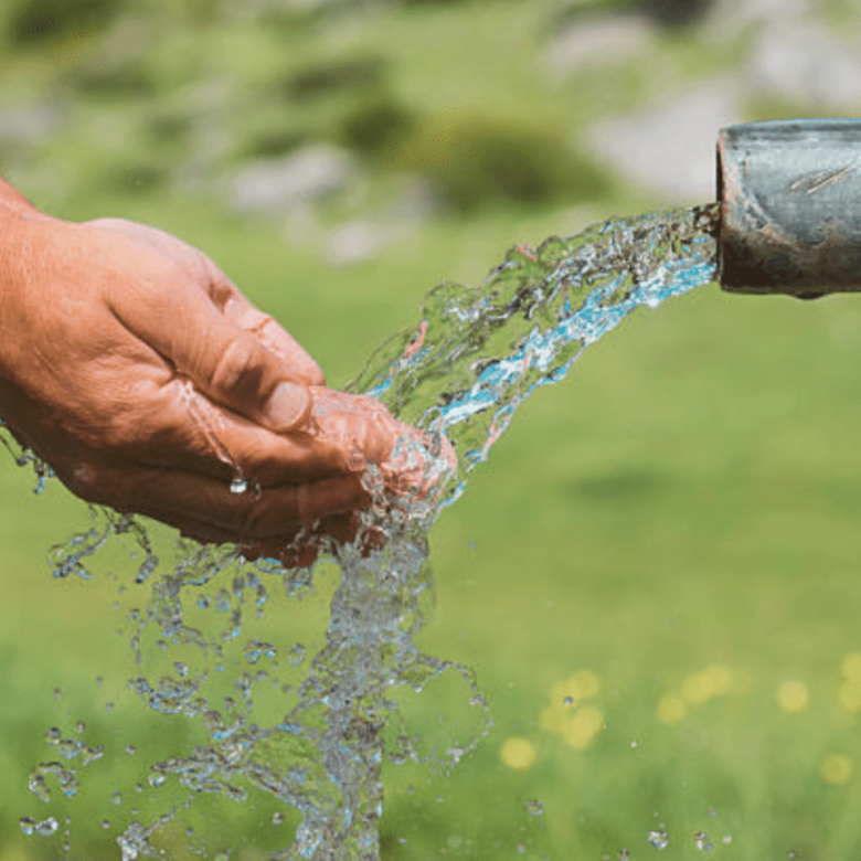 Hands collecting clean water flowing from a metal pipe with a green meadow in the background.