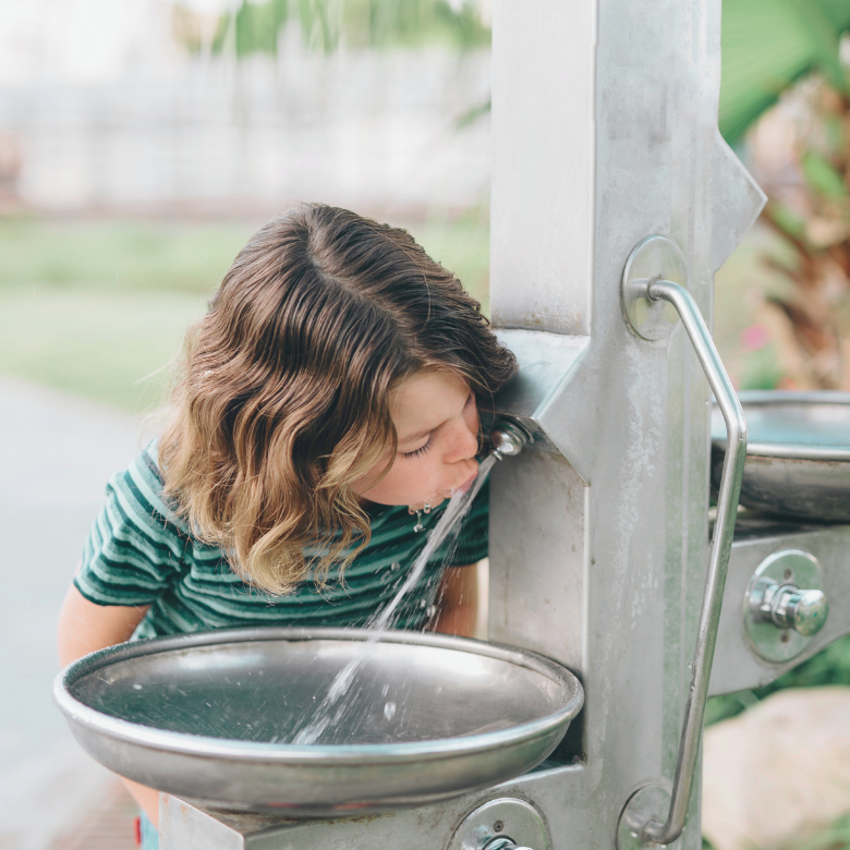 A child drinking water from a public stainless steel drinking fountain outdoors.