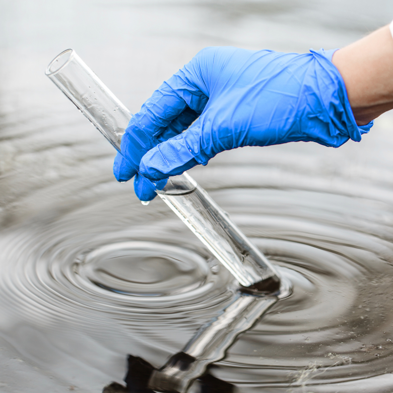 A person wearing a blue protective glove collects a water sample into a glass test tube from a dark water body.
