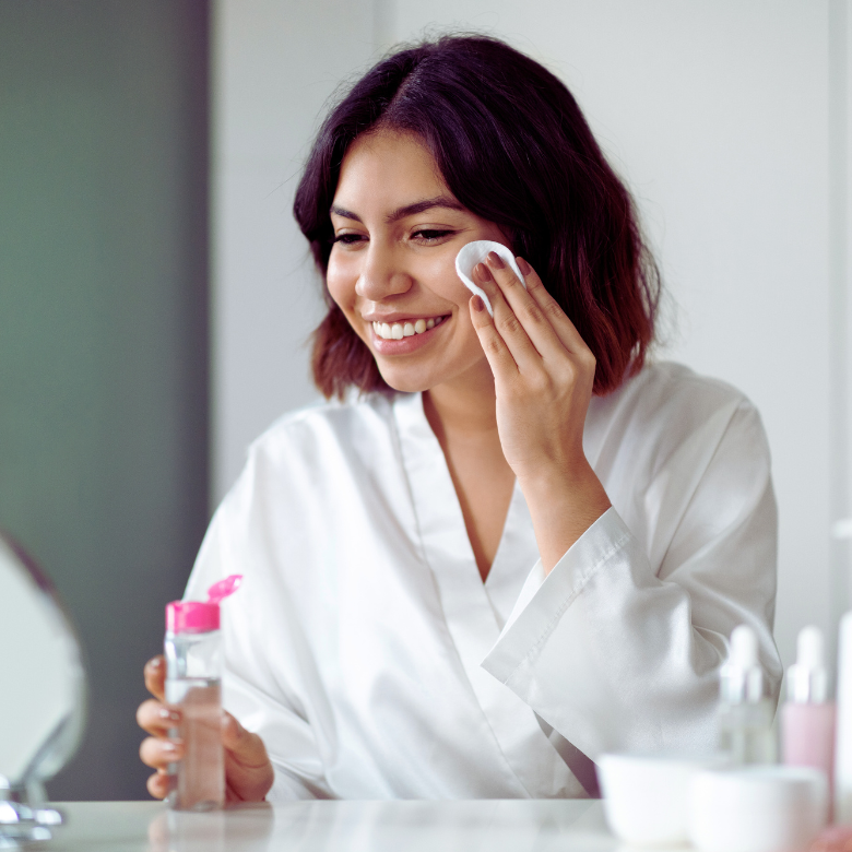 Seorang wanita berjubah putih tersenyum sambil menyapu toner pada mukanya dengan kapas di hadapan cermin.