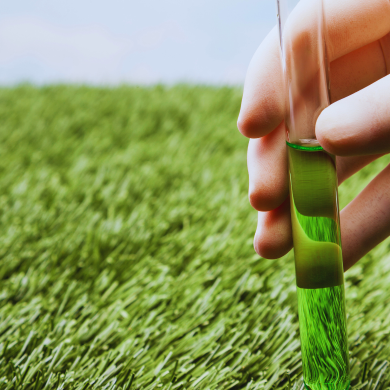 Una mano sostiene un tubo de ensayo con líquido verde sobre un fondo de campo de cultivo.