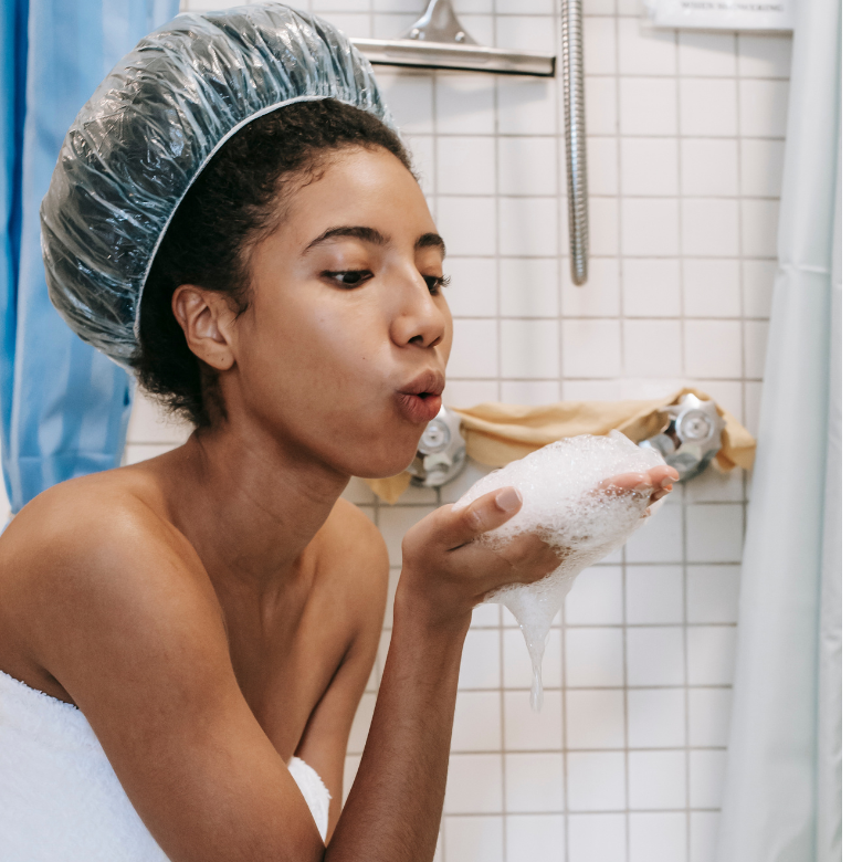 Duş başlığı takmış bir kadın, duşta elinde tuttuğu köpüğe üflüyor.